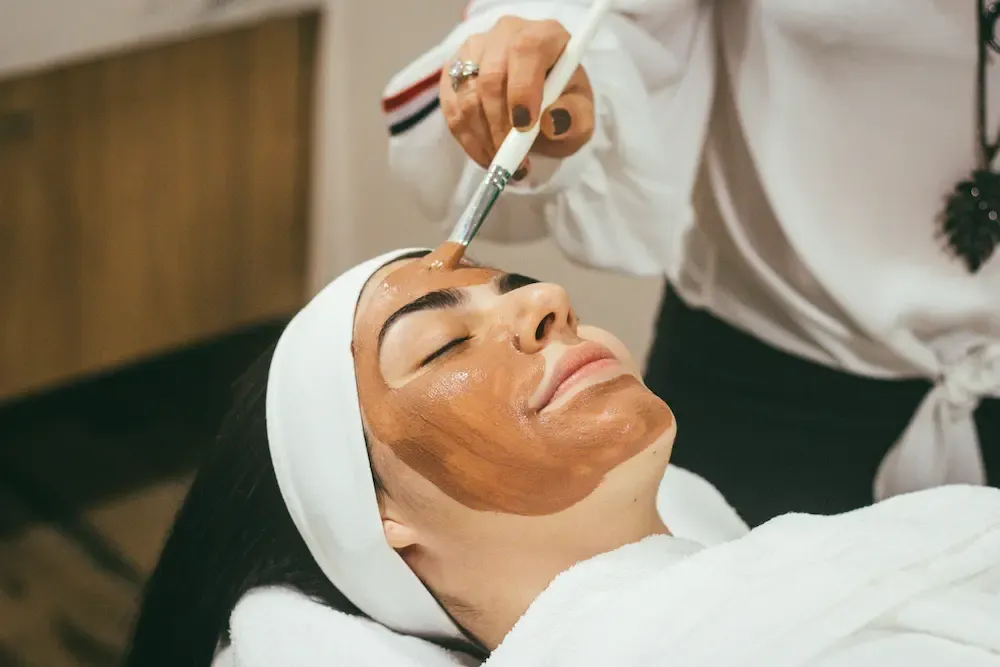 A woman with a towel on her head having her face painted with a brush-applied mask
