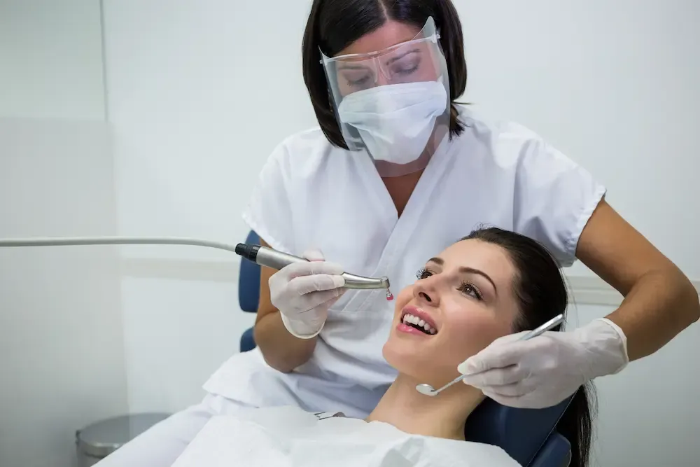 Dentist in face mask and protective gear performing a professional dental cleaning on a female patient.