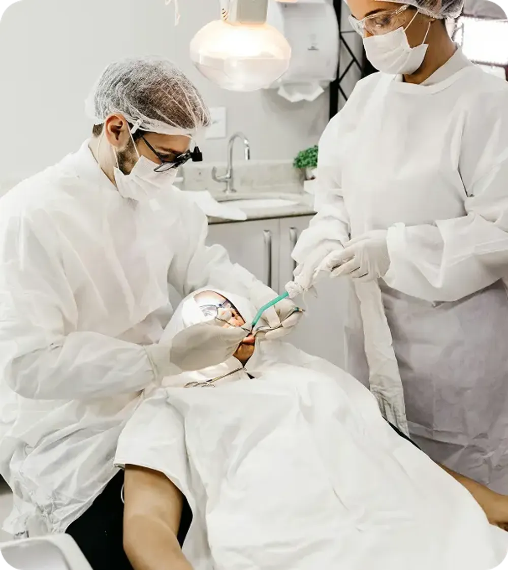 Two professional dentists in white scrubs, face masks, and hair caps performing a precision dental procedure on a patient.