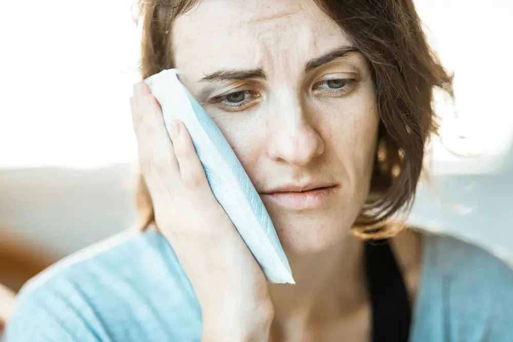 Woman in pain holding a tissue to her swollen cheek, needing urgent dental emergency care.