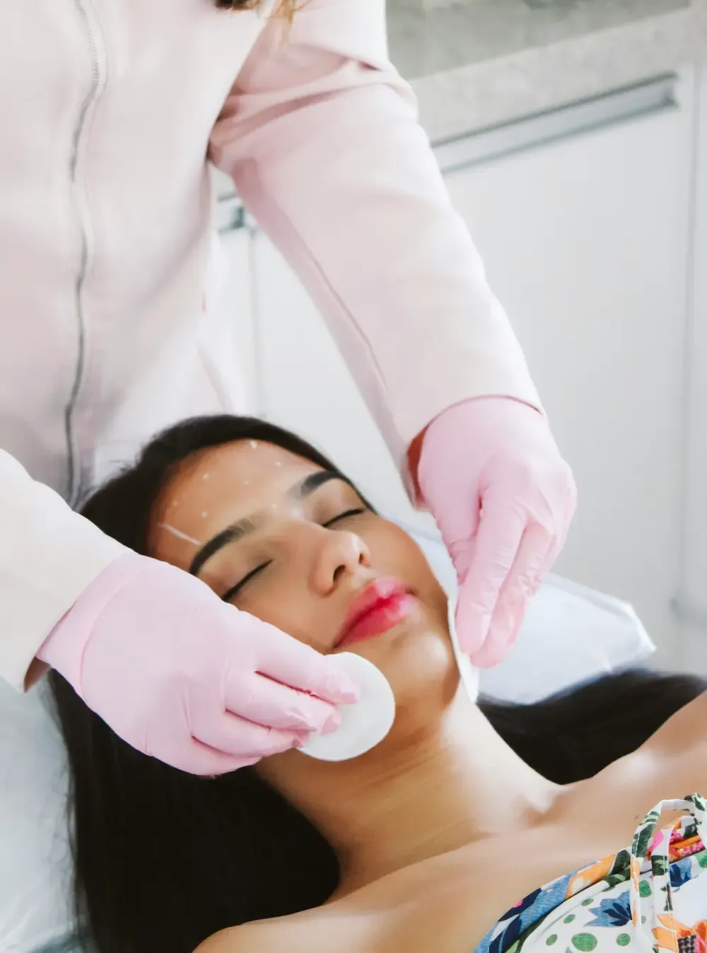 A doctor wearing pink gloves applying two round cotton pads on a brunette woman’s face